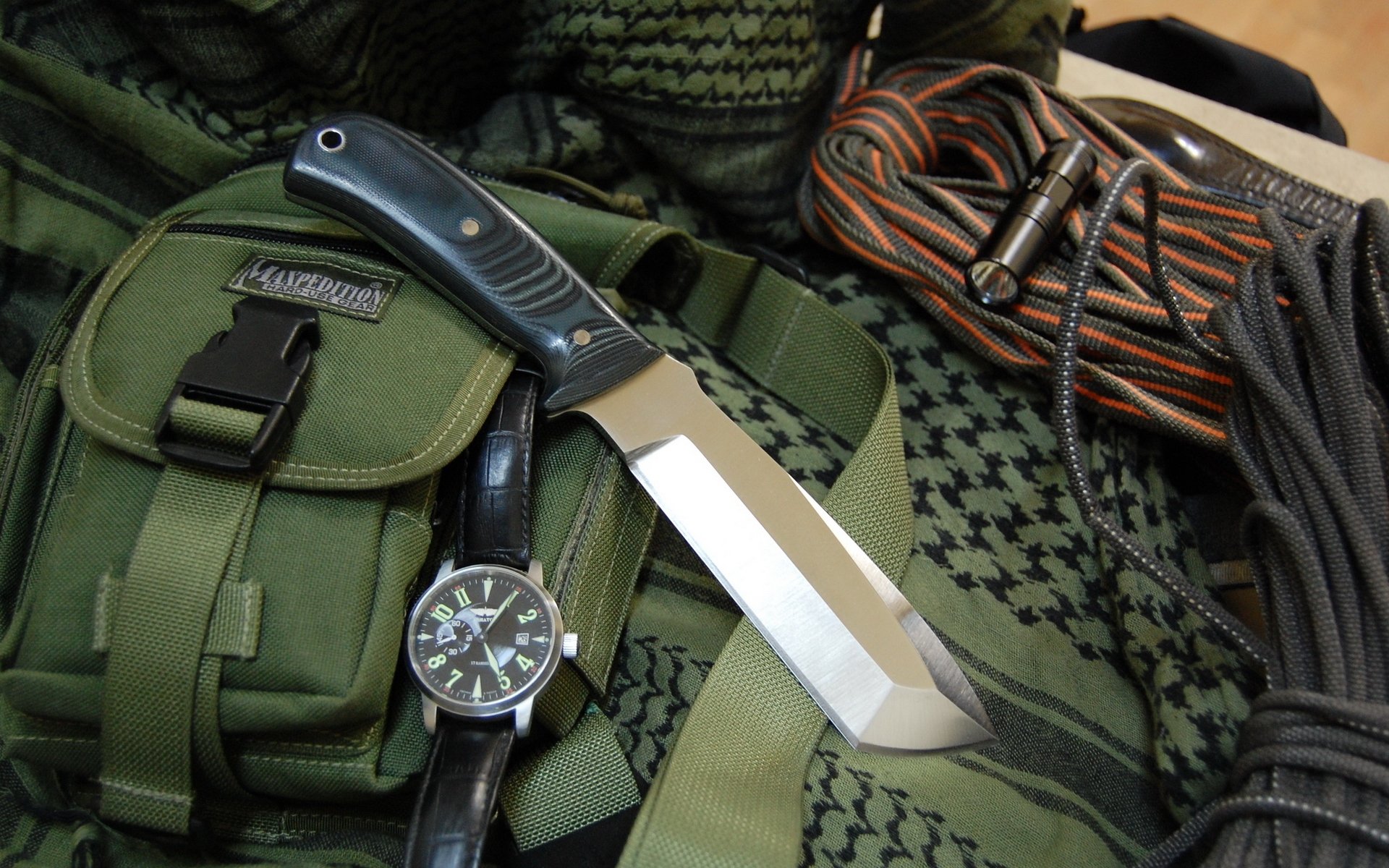A high-definition PC desktop wallpaper featuring a tactical knife resting on a green backpack alongside a watch and climbing rope, evoking an adventurous and rugged aesthetic.