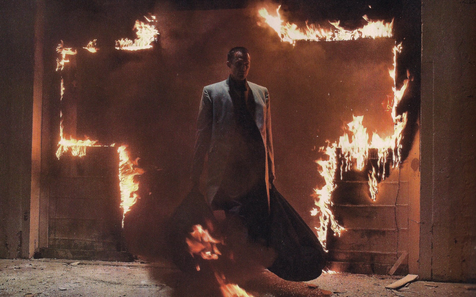 2K Quad HD PC desktop wallpaper from the movie Legion: lone man in a long coat steps through a doorway framed by roaring flames in a dark, smoky room.