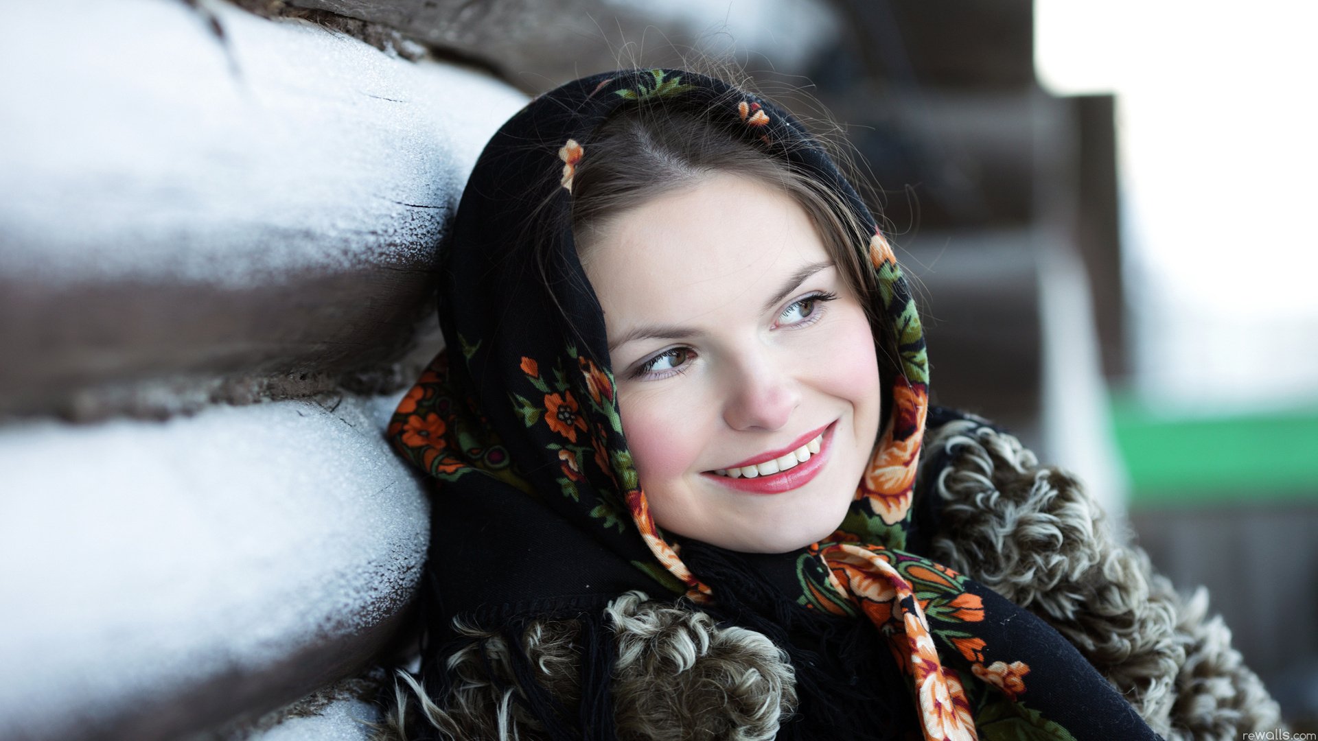 HD desktop wallpaper featuring a pretty woman smiling warmly, wearing a traditional floral headscarf and fur-lined coat, leaning against a snow-covered wooden surface.