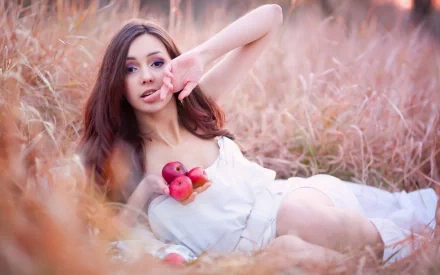 HD wallpaper of a beautiful woman in a white dress lying in a field, holding apples with gentle light creating a serene background.