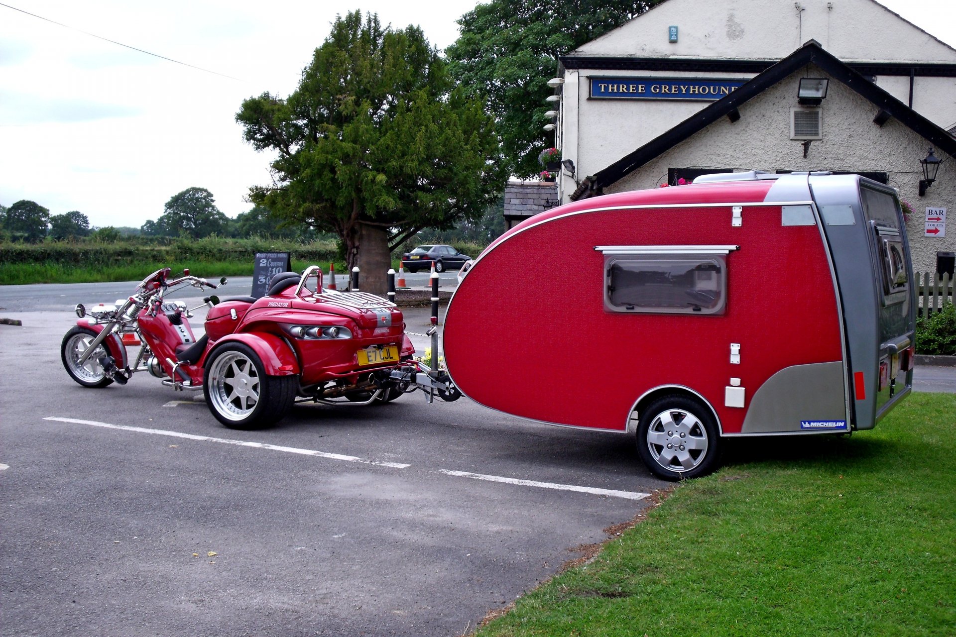 HD PC desktop wallpaper featuring a red motorcycle towing a matching red teardrop camper in a parking lot near a countryside building.
