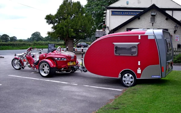 HD PC desktop wallpaper featuring a red motorcycle towing a matching red teardrop camper in a parking lot near a countryside building.