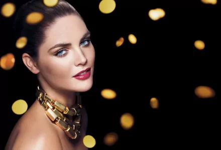 Hilary Rhoda with striking blue eyes and red lipstick, wearing a bold gold necklace, set against a blurred dark background with glowing bokeh lights in 4K Ultra HD.