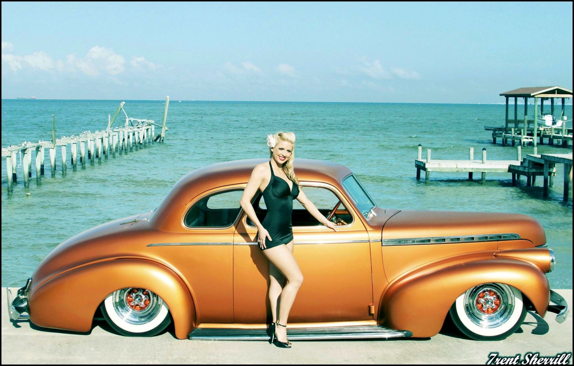 A stylish woman poses beside a custom Chevrolet lowrider against a vibrant seaside backdrop, showcasing the fusion of automotive culture and beauty.