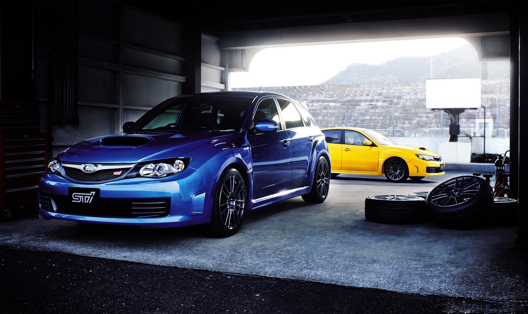 HD PC desktop wallpaper background showing two Subaru vehicles—a blue STI and a yellow hatch—parked in a dim garage with a city view.