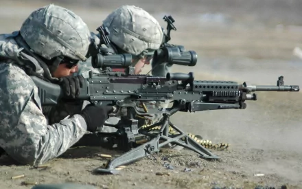 HD desktop wallpaper featuring two soldiers in military gear aiming a machine gun, set in a dusty outdoor environment.