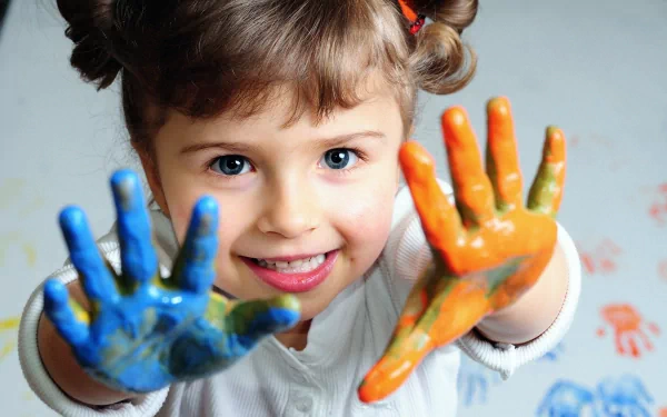 A cute child joyfully displays her paint-covered hands, showcasing vibrant colors. This HD image serves as a delightful desktop wallpaper, capturing the essence of creativity and fun.