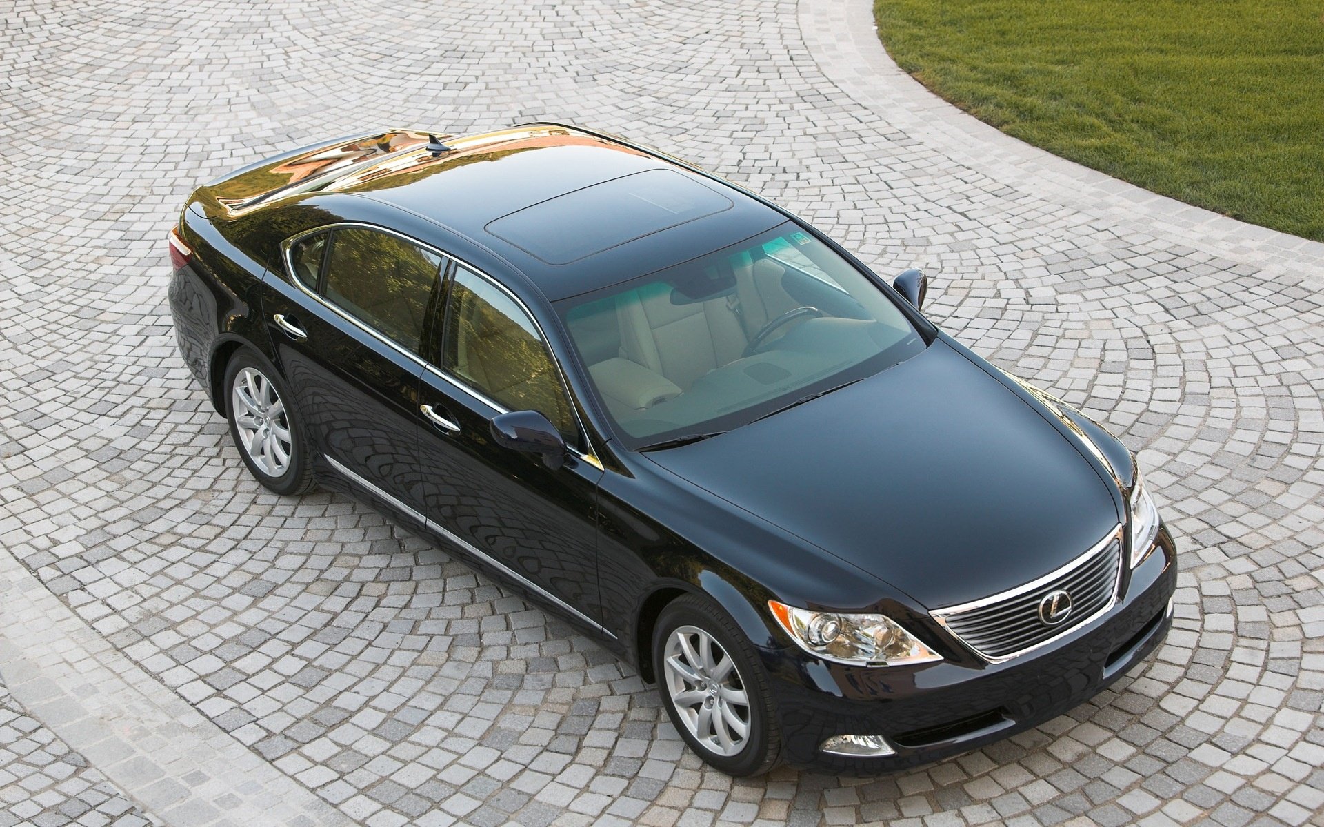 HD PC desktop wallpaper/background showing a black Lexus sedan parked on a curved cobblestone driveway, high-angle view of the vehicle with a patch of green lawn in the background.
