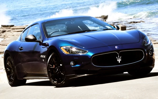 HD PC desktop wallpaper featuring a sleek blue Maserati vehicle parked by a coastal road with waves and rocks in the background.