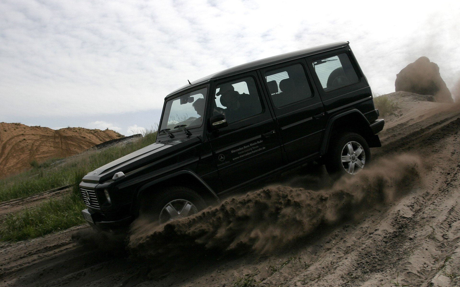 HD PC desktop wallpaper background of a Mercedes vehicle driving off-road, kicking up sand along a rugged trail beneath a cloudy sky