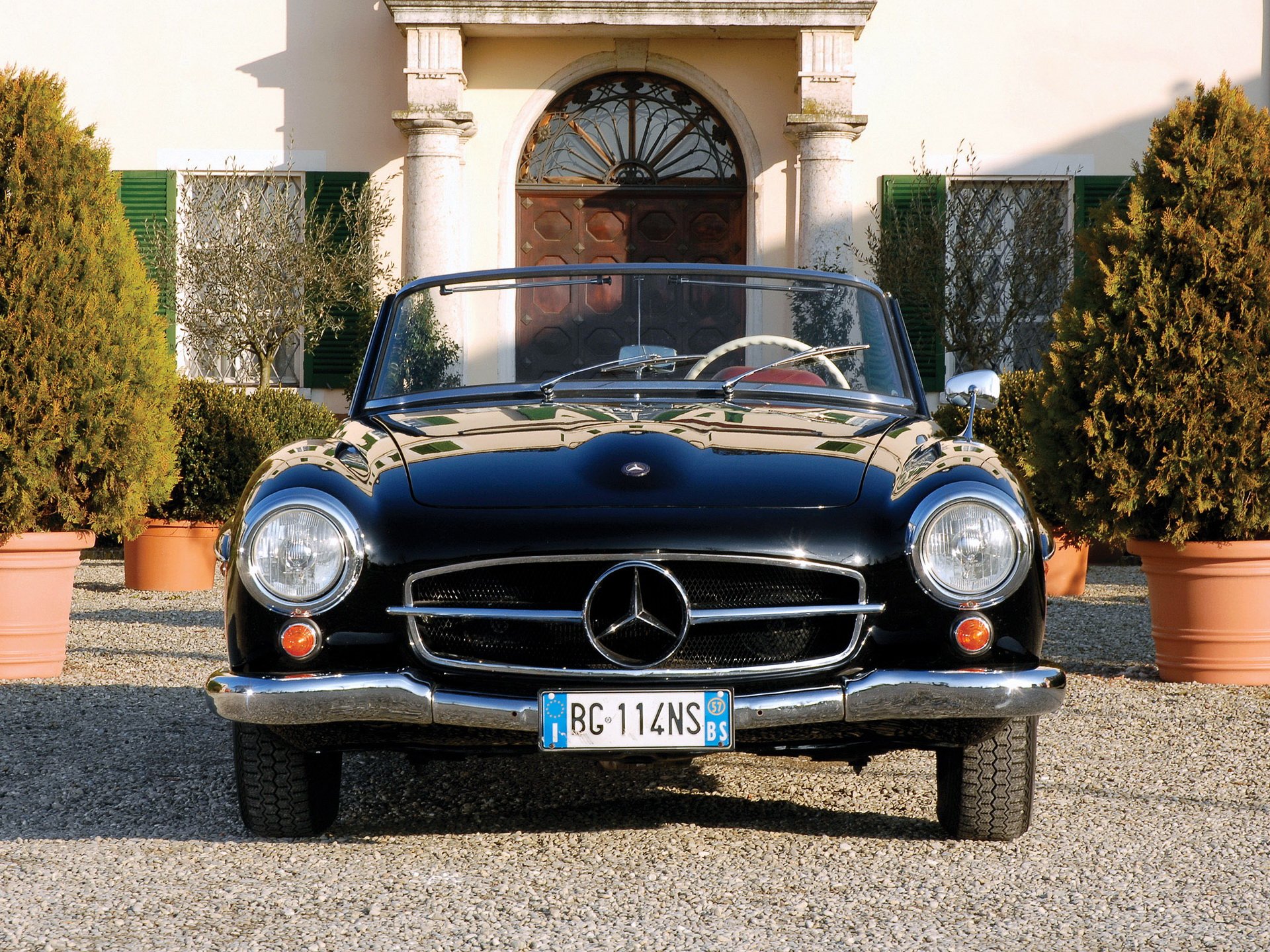 HD PC desktop wallpaper showing a classic black Mercedes convertible vehicle, front view parked before a villa entrance flanked by potted trees.