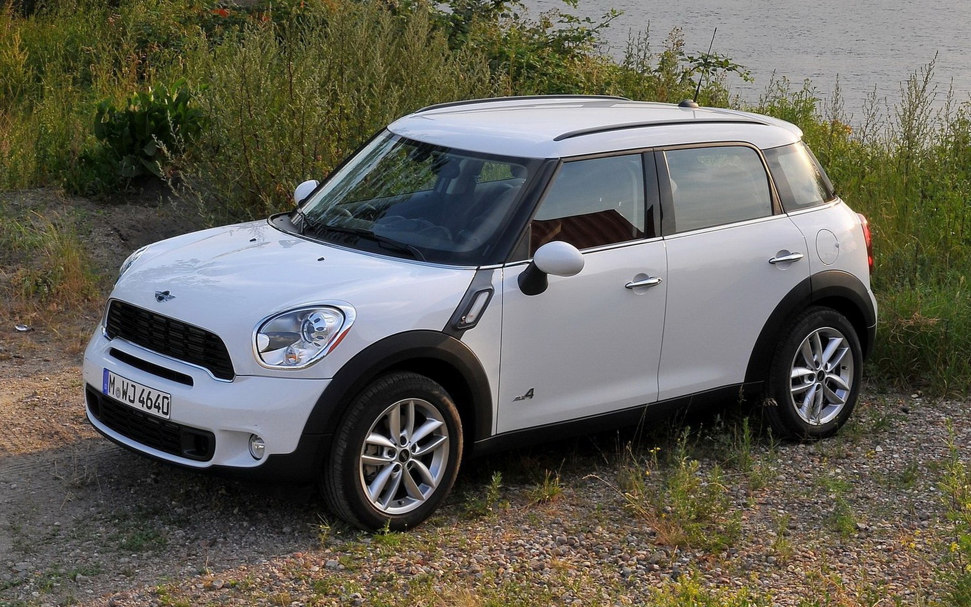 White Mini Cooper vehicle parked on gravel near grass and water, three-quarter front view — HD PC desktop wallpaper and background of a compact car.