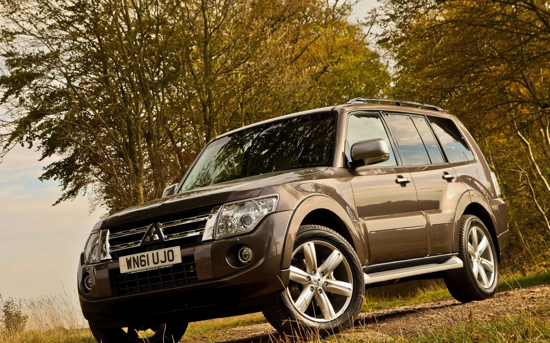 HD PC desktop wallpaper featuring a brown Mitsubishi SUV parked on a grassy area with autumn trees in the background.