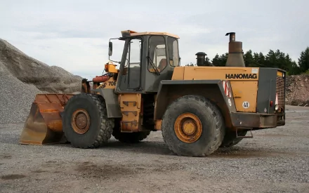 HD desktop wallpaper featuring a Hanomag 70E wheel loader vehicle positioned on a gravel surface with a cloudy sky backdrop.