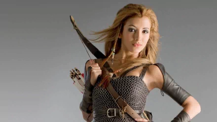 A woman warrior, inspired by India de Beaufort's role in Kröd Mändoon and the Flaming Sword of Fire, poses confidently with a bow, set against a sleek gray background.