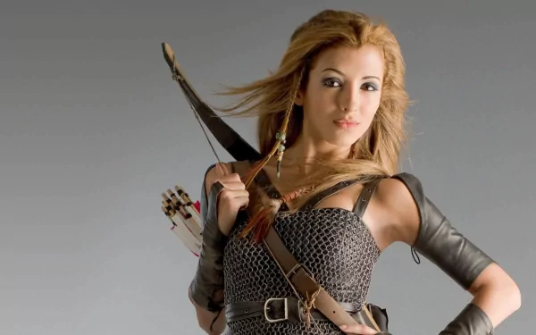 A woman warrior, inspired by India de Beaufort's role in Kröd Mändoon and the Flaming Sword of Fire, poses confidently with a bow, set against a sleek gray background.