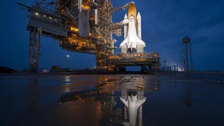 The image features NASA's Space Shuttle Atlantis, illuminated and poised for launch at twilight, with its reflection visible on the wet ground, creating a striking HD wallpaper.