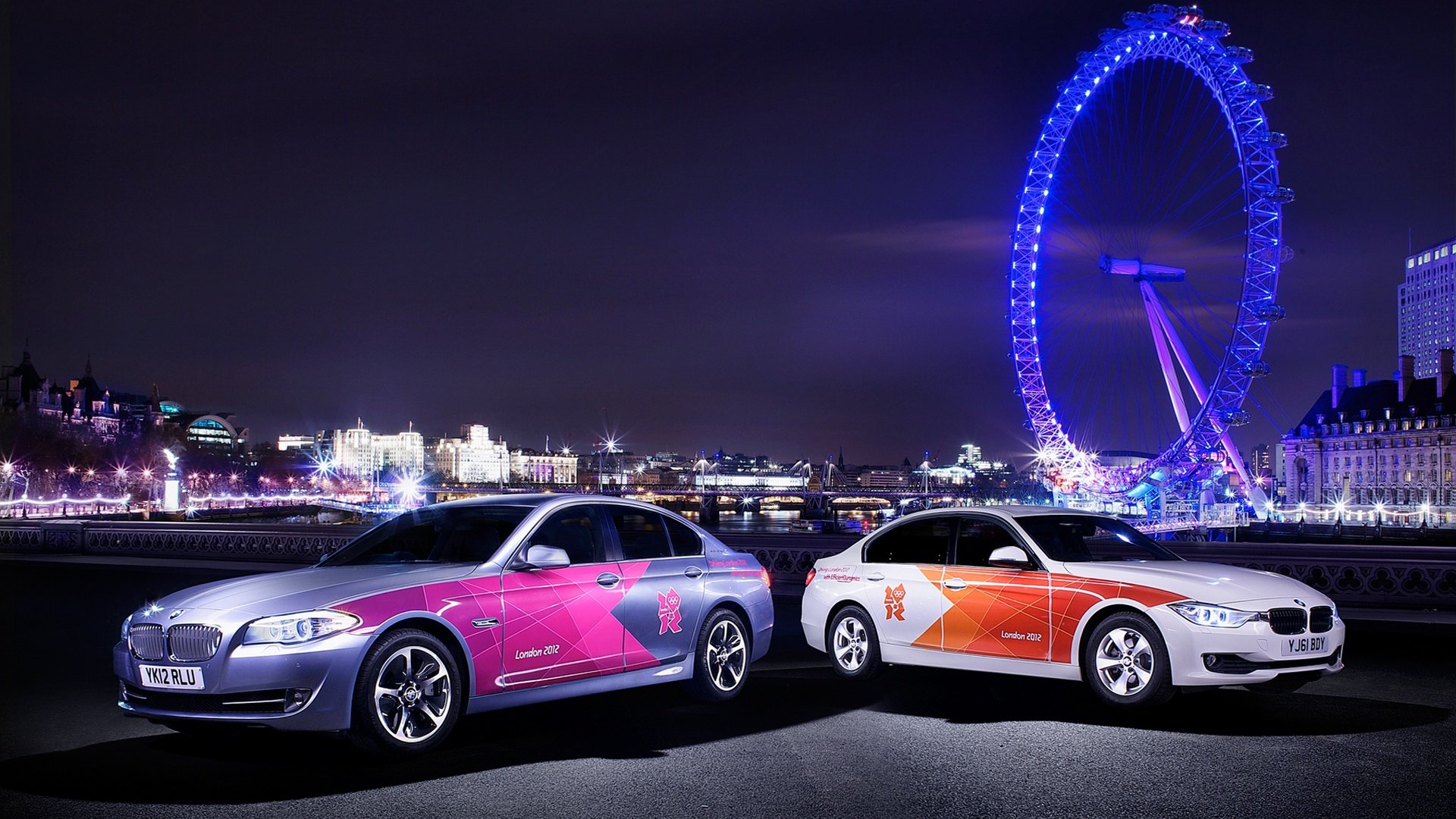 Two BMW cars with vibrant decals are parked at night with a brightly lit Ferris wheel in the background, capturing a dynamic scene linked to the Olympic Games.