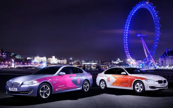 Two BMW cars with vibrant decals are parked at night with a brightly lit Ferris wheel in the background, capturing a dynamic scene linked to the Olympic Games.