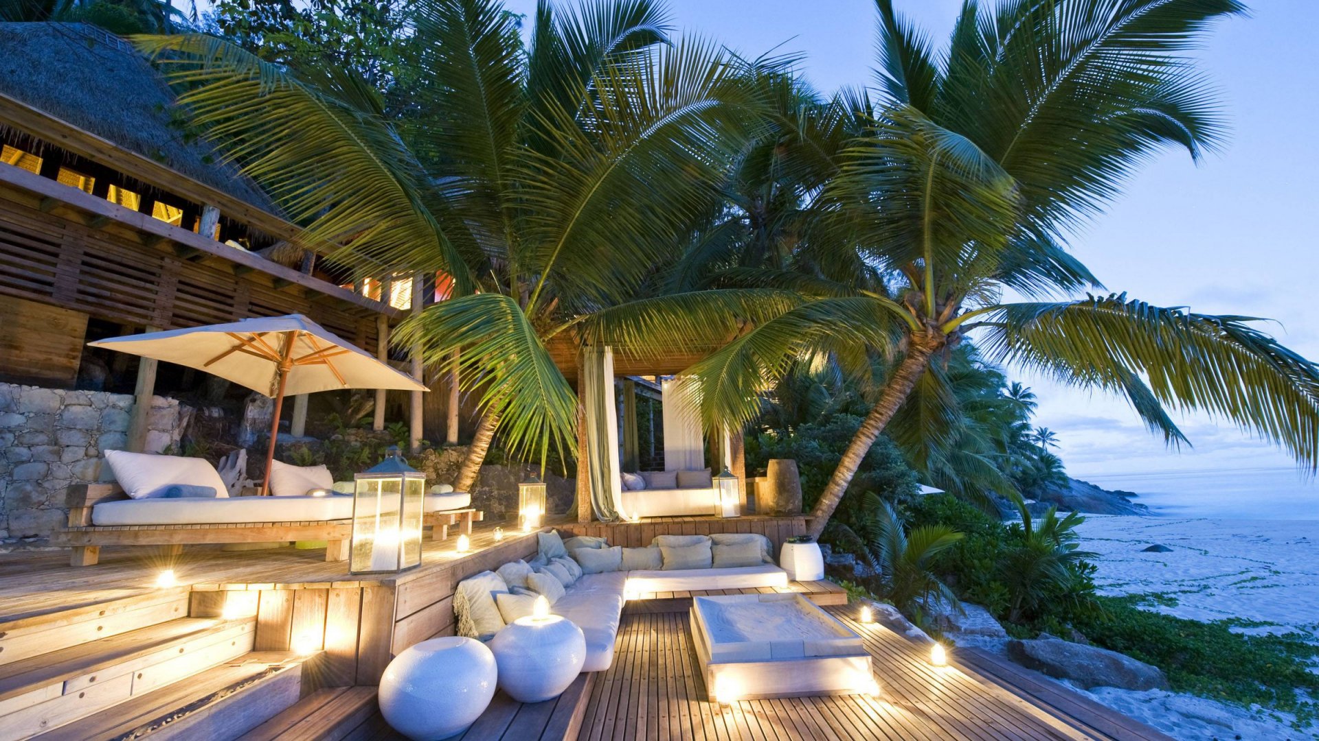 A serene tropical resort in the Seychelles, featuring lush palm trees and a cozy outdoor seating area, illuminated by soft lights against a tranquil ocean backdrop.