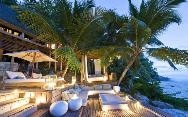 A serene tropical resort in the Seychelles, featuring lush palm trees and a cozy outdoor seating area, illuminated by soft lights against a tranquil ocean backdrop.