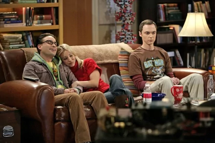HD desktop wallpaper of The Big Bang Theory cast seated on a cozy living-room couch — two men and a woman relaxed amid bookshelves, coffee table and sitcom set props.