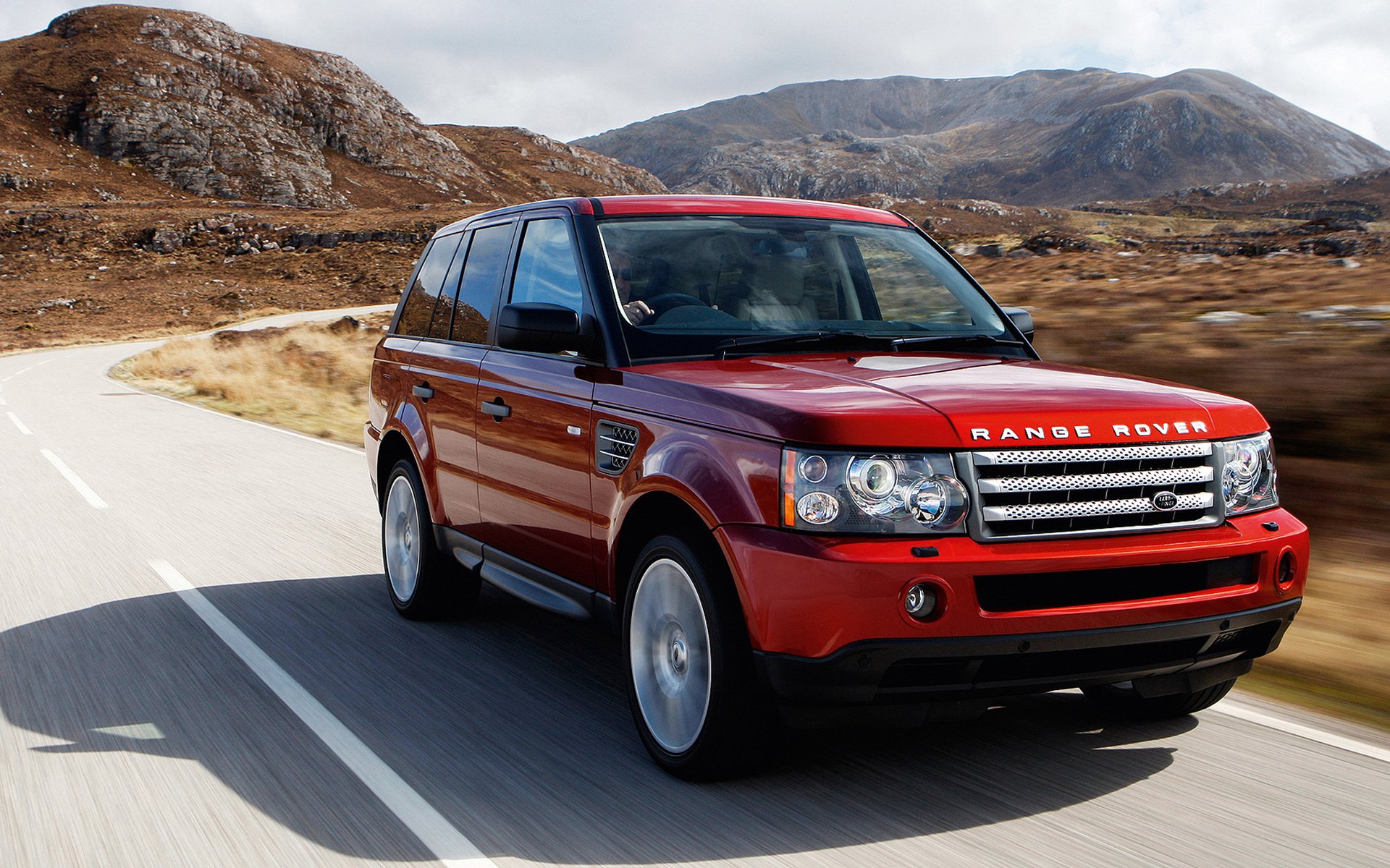 Red Range Rover driving on a mountain road, captured in an HD PC desktop wallpaper background with rugged terrain and cloudy sky.