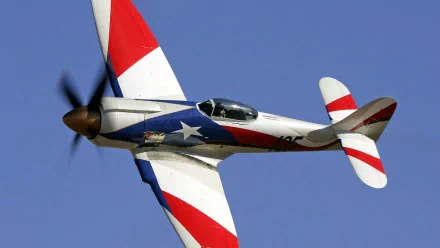 HD PC desktop wallpaper background of a red-white-blue military aircraft with spinning propeller soaring across a clear blue sky.