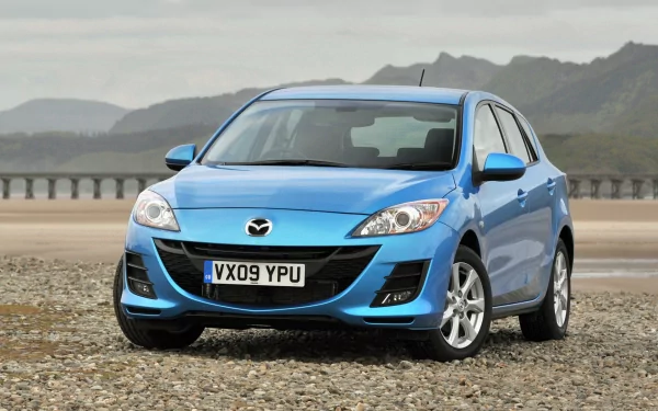 HD PC desktop wallpaper of a blue Mazda hatchback on a pebble beach with distant mountains and a cloudy sky.