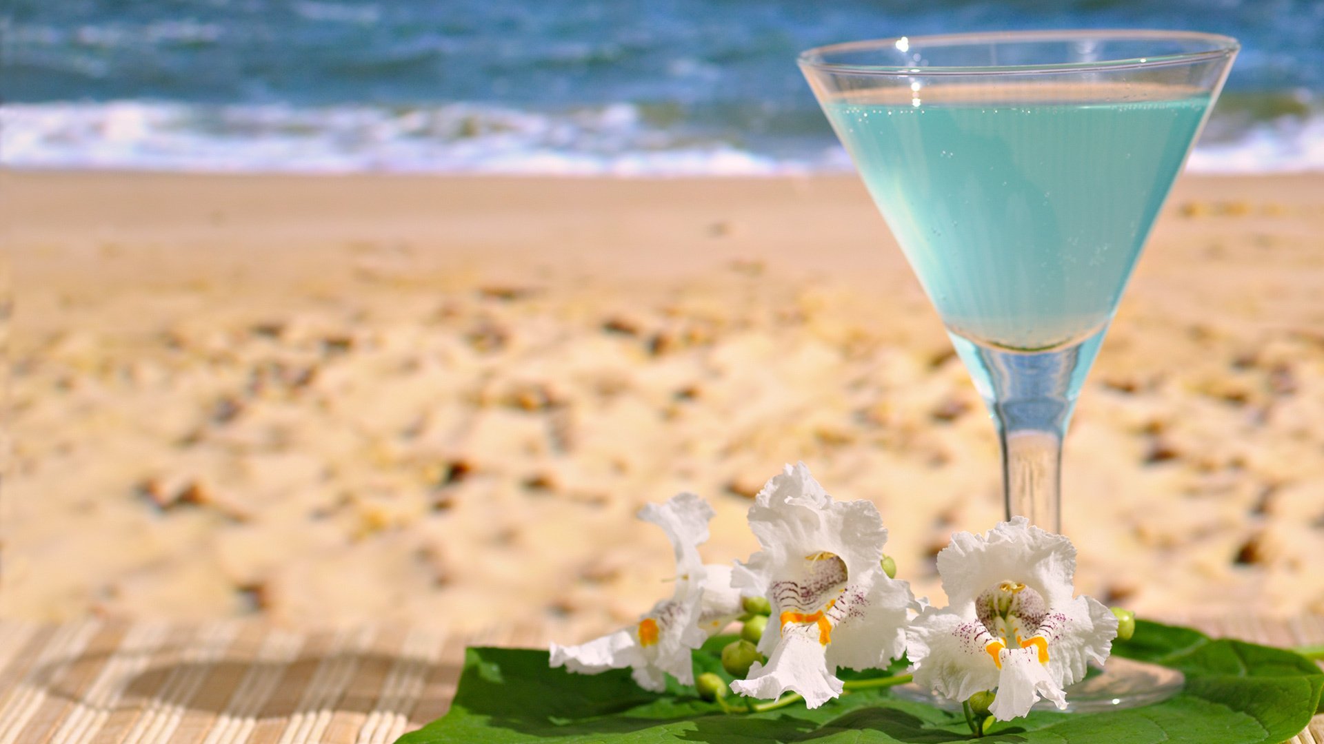 Pastel blue cocktail in a glass on a green leaf with white flowers, set against a sandy beach and ocean background, captured in HD for a desktop wallpaper.