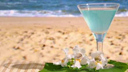 Pastel blue cocktail in a glass on a green leaf with white flowers, set against a sandy beach and ocean background, captured in HD for a desktop wallpaper.