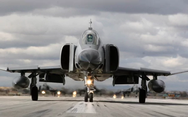 A McDonnell Douglas F-4 Phantom II military jet approaches the runway, surrounded by dramatic clouds, showcasing its sleek design and power in this HD desktop wallpaper.