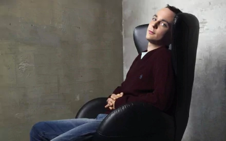 HD desktop wallpaper featuring Jim Parsons as Sheldon Cooper from the TV show The Big Bang Theory, seated in a black chair against a textured concrete wall.