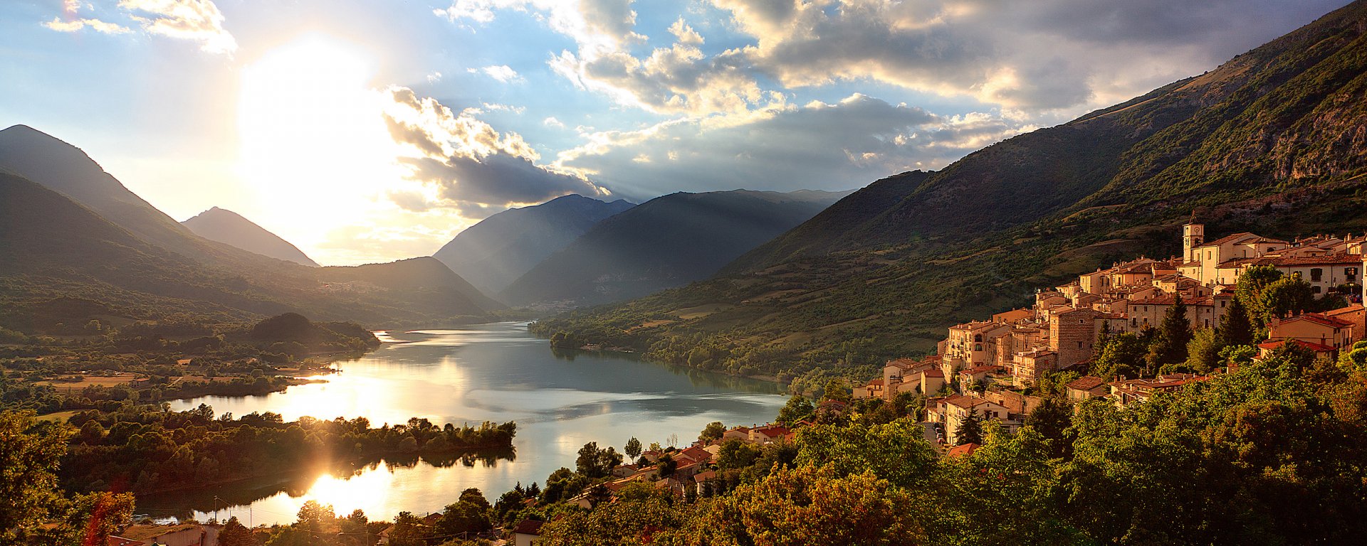 HD desktop wallpaper featuring a scenic view of Lake Barrea, Italy, with sunlit clouds, mountains, and a lakeside village surrounded by greenery.