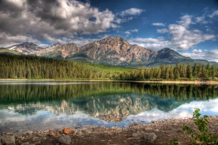 Breathtaking view of the Canadian Rockies, showcasing majestic mountains reflecting in a serene lake surrounded by lush forests under a dynamic sky. Ideal nature backdrop for a desktop wallpaper.