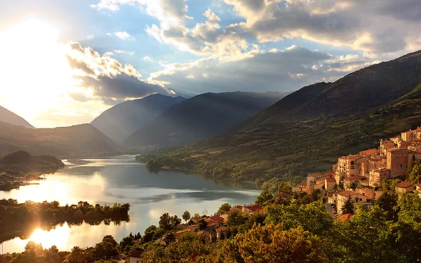 HD desktop wallpaper featuring a scenic view of Lake Barrea, Italy, with sunlit clouds, mountains, and a lakeside village surrounded by greenery.
