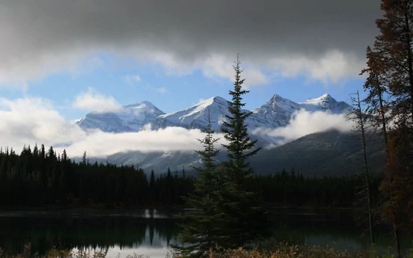 Clouds hover above the rocky mountains of Canada, reflecting over a calm lake surrounded by dense forest in this 4K Ultra HD nature desktop wallpaper.