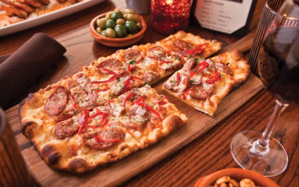HD desktop wallpaper of a delicious rectangular pizza topped with pepperoni and red peppers, served on a wooden board alongside olives, nuts, and a glass of red wine.