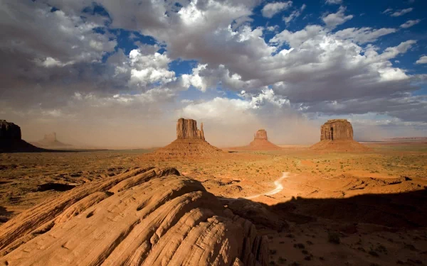 A scenic HD desktop wallpaper capturing a winding path through the desert landscape of Monument Valley under a dramatic cloud-filled sky.