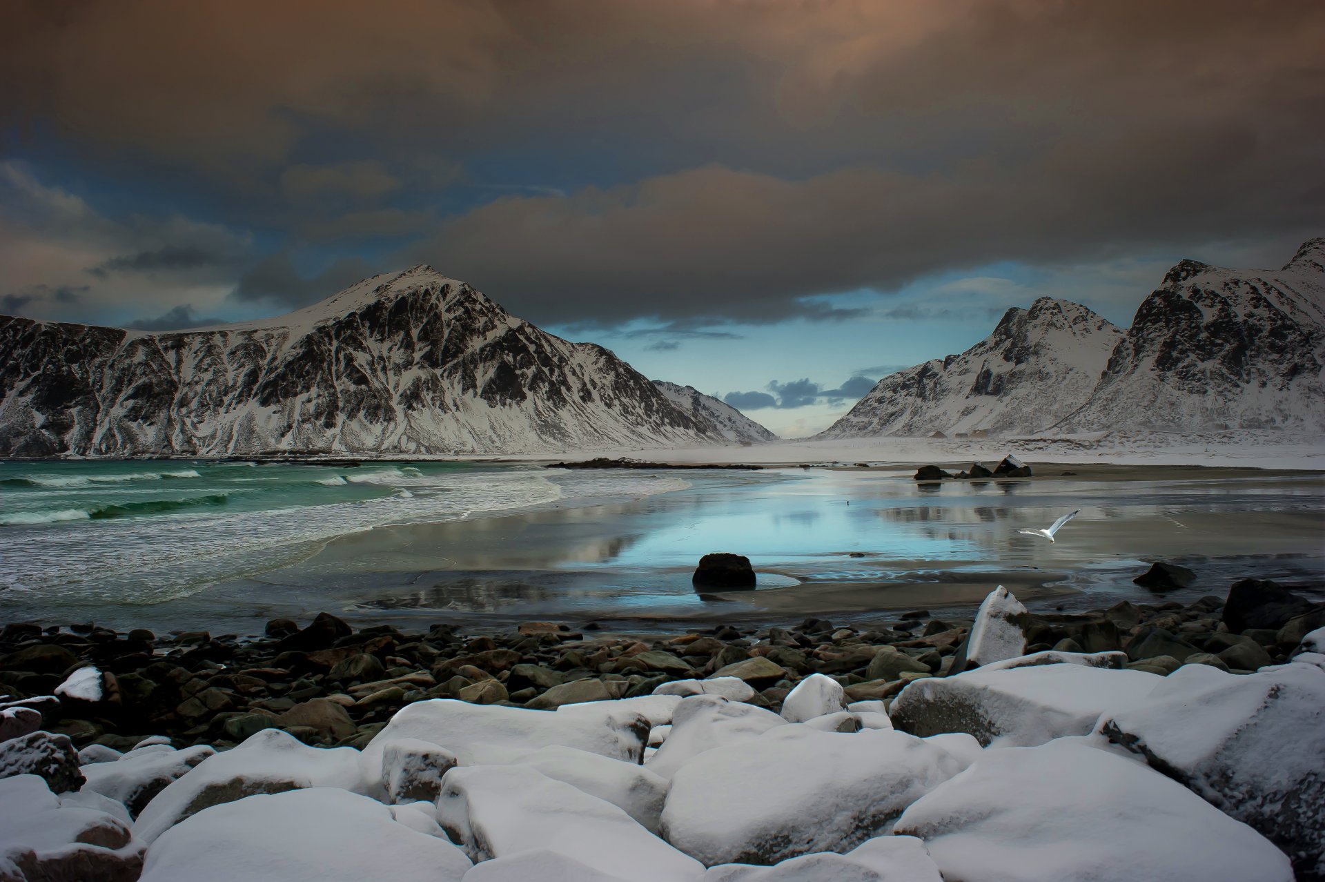 Snow-covered mountains reflected in a serene, icy lake with scattered rocks in the foreground under a cloudy sky. This HD nature landscape makes an ideal desktop wallpaper or background.