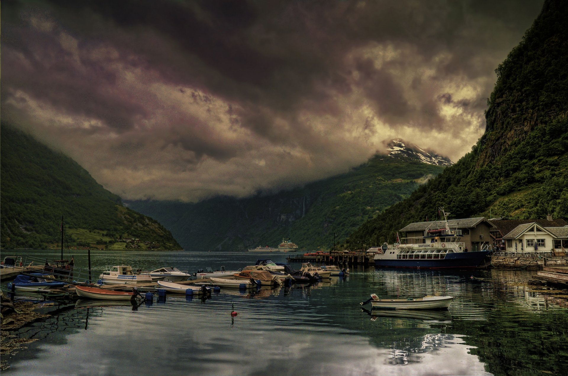 HD desktop wallpaper featuring a serene man-made town with boats docked along calm waters, surrounded by mountains under a dramatic cloudy sky.