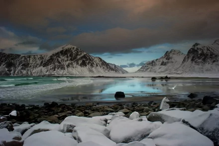 Snow-covered mountains reflected in a serene, icy lake with scattered rocks in the foreground under a cloudy sky. This HD nature landscape makes an ideal desktop wallpaper or background.