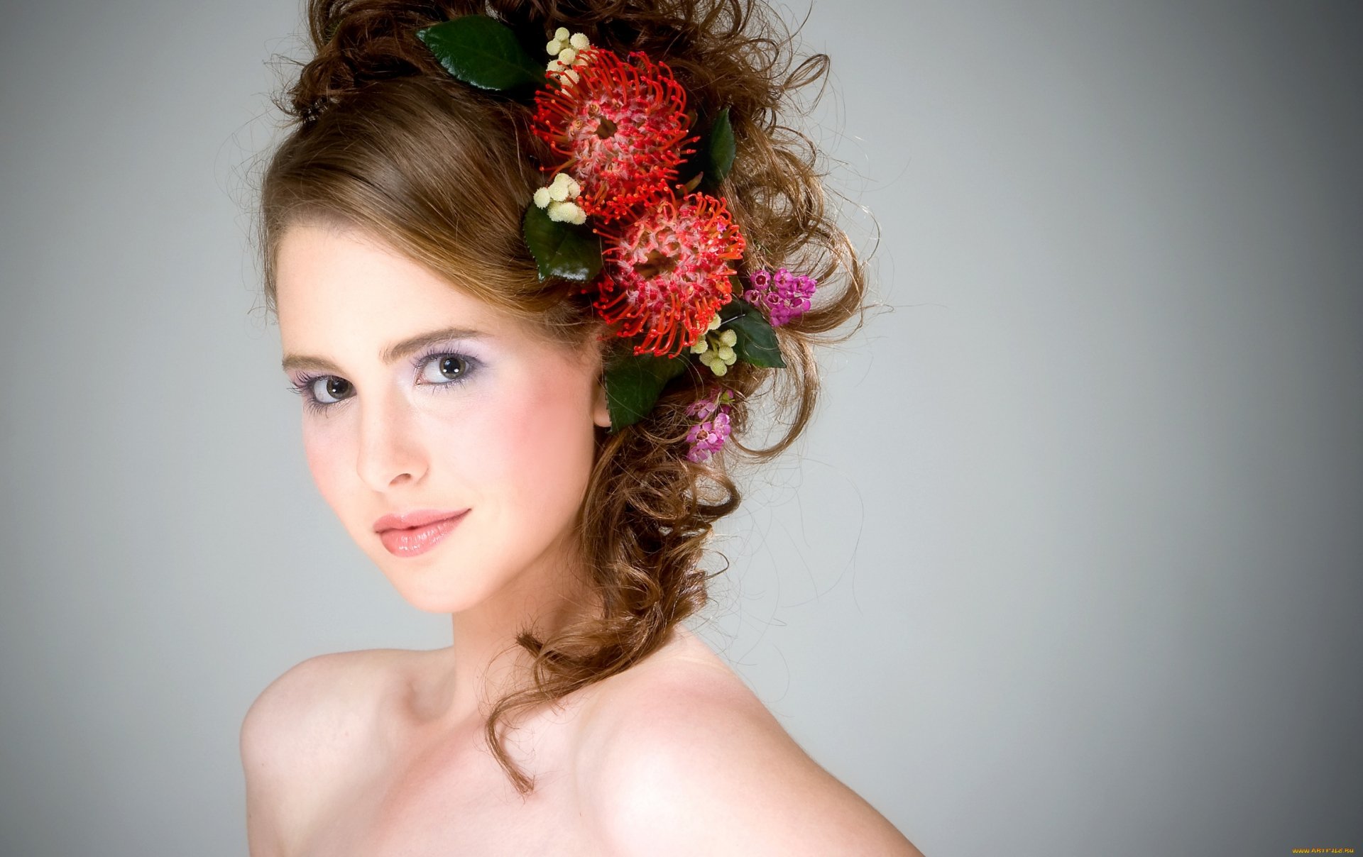 A model with voluminous, curly hair adorned with vibrant flowers poses confidently against a soft gray background, showcasing a striking and artistic hairstyle.