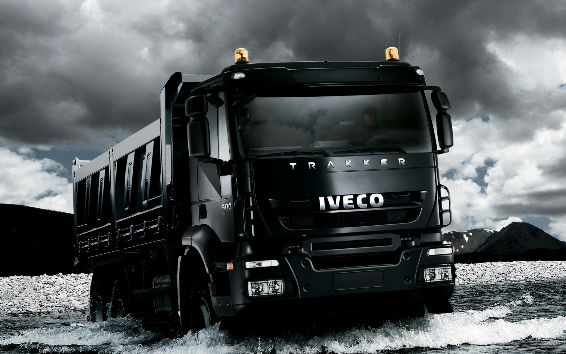 HD PC desktop wallpaper of an Iveco vehicle: black dump truck splashing through shallow water beneath dramatic storm clouds, high-contrast automotive background.