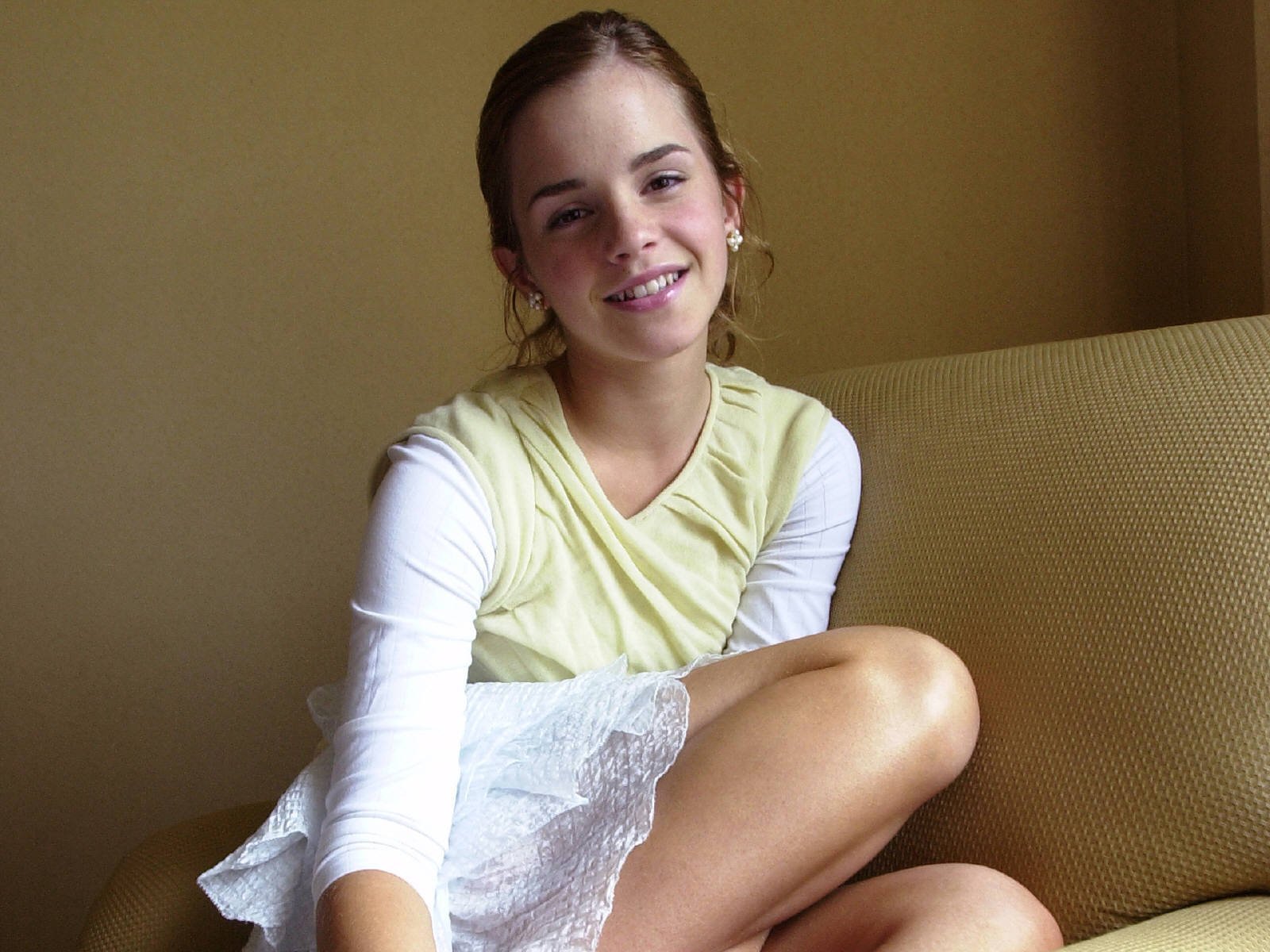 A cheerful Emma Watson sits on a couch, dressed in a light yellow top and white lace skirt, showcasing a natural smile in this high-definition desktop wallpaper.