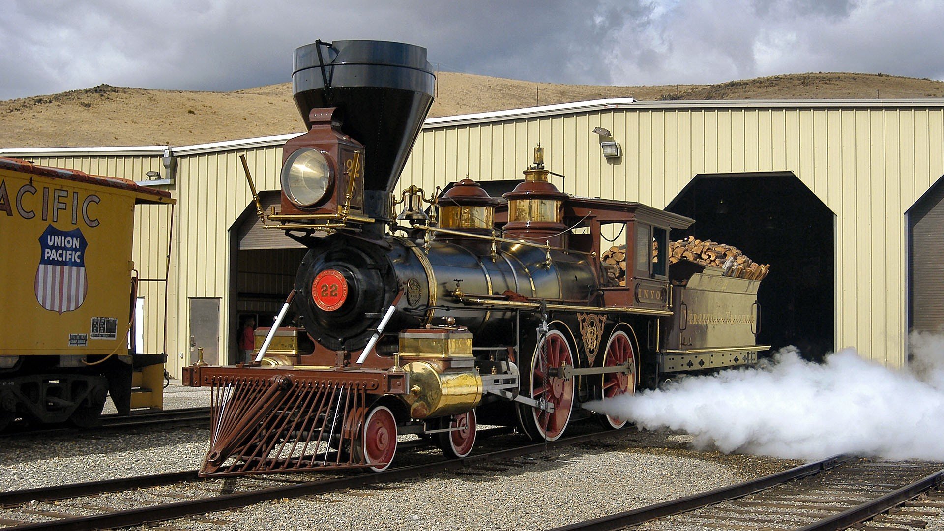 A vintage steam locomotive with brass and red details emits steam as it pauses near a cream-colored industrial building, captured in HD for a PC desktop wallpaper background.