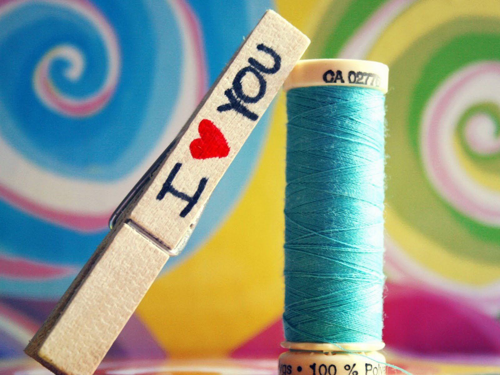 A vibrant HD wallpaper featuring a clothespin marked I ♥ YOU and a spool of turquoise thread, set against a colorful swirling background that captures the essence of love.