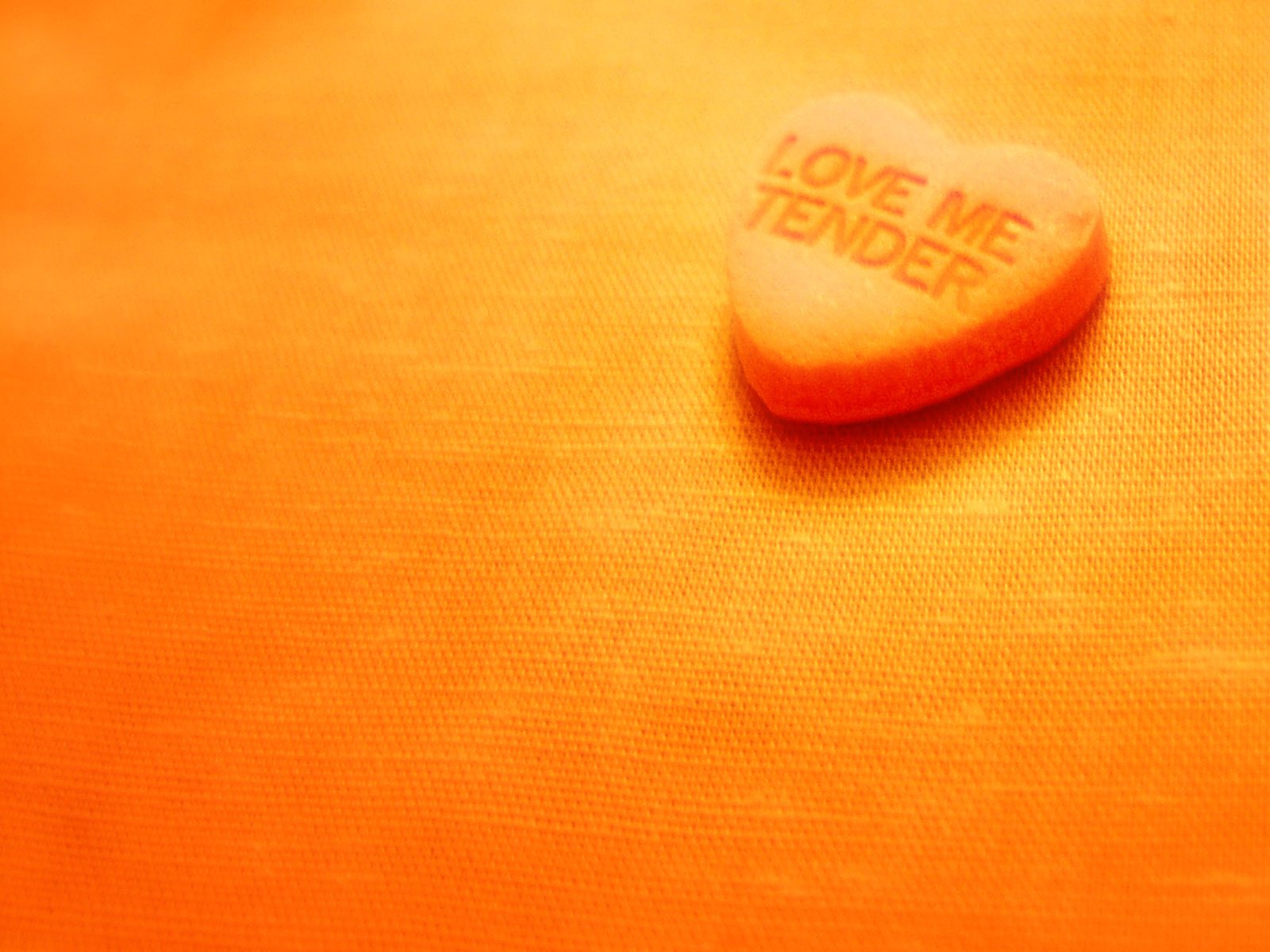 A vibrant HD desktop wallpaper featuring an artistic depiction of a heart-shaped candy with the words LOVE ME TENDER on a warm orange background, celebrating love and affection.