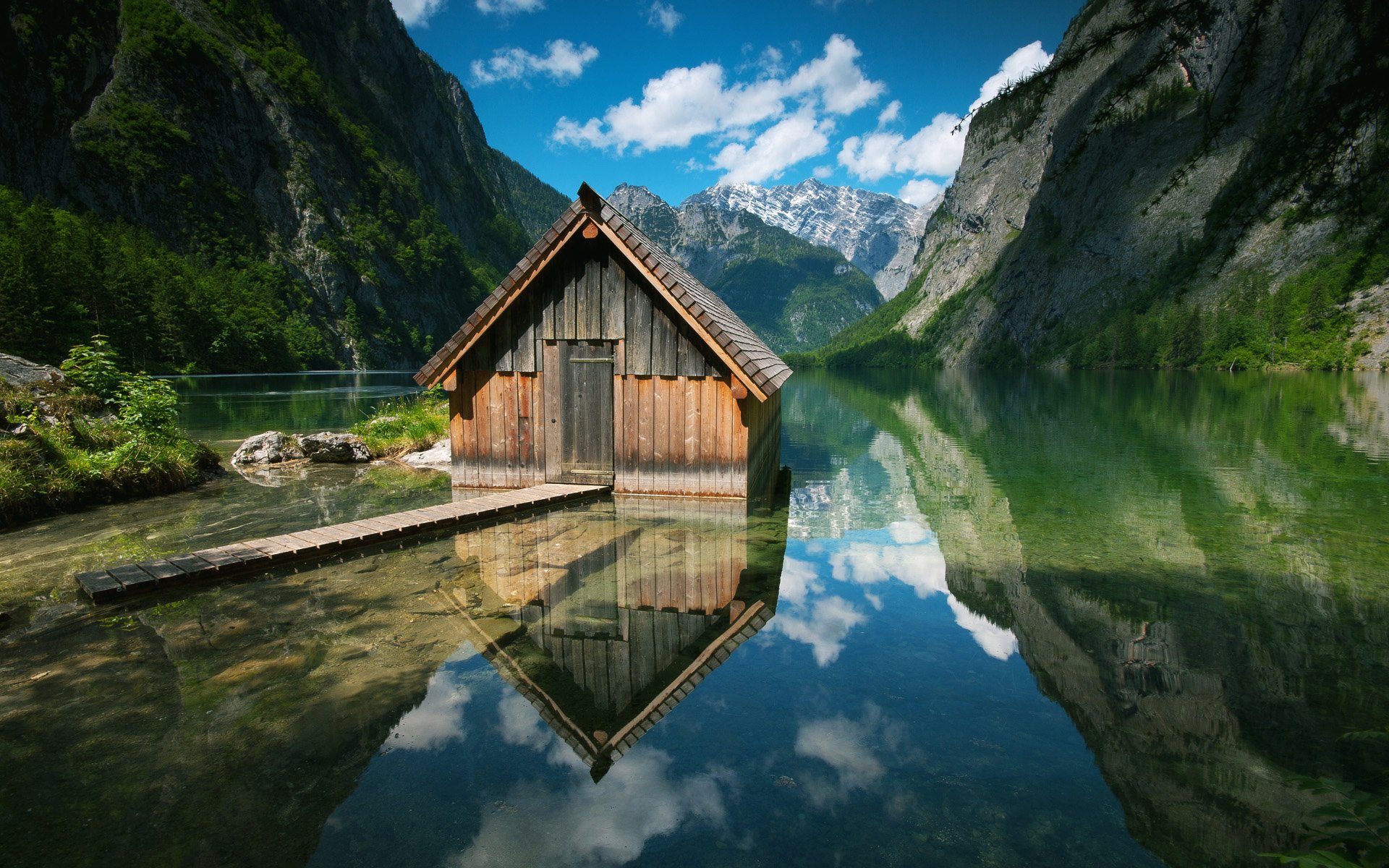 A charming wooden hut by a serene lake, surrounded by majestic mountains in Germany, reflects beautifully in the clear water under a bright blue sky. Captivating HD photography.
