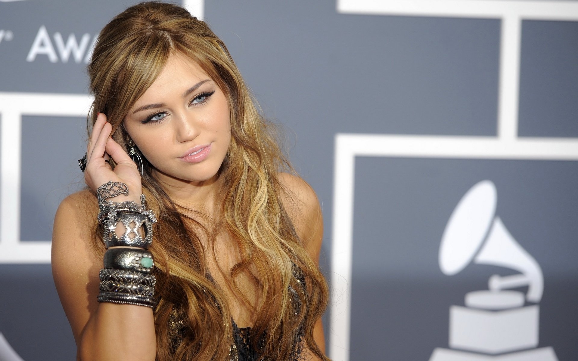 HD PC desktop wallpaper of a female pop singer at a music awards backdrop, long wavy hair and stacked bracelets, posing thoughtfully as a music-themed celebrity portrait.